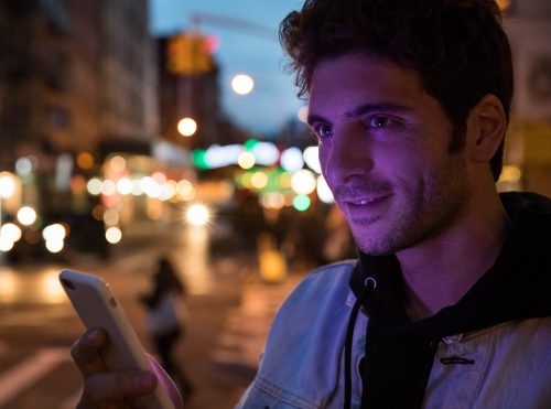 A man is lit by the light of his phone screen as he stands on a busy city street.