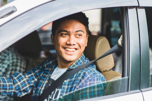 A photo of a young Uber driver smiling out the window of his silver car.