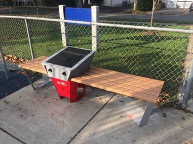 Image is of a small grill with two wooden tables sticking out of both sides of it. The grill is outside.