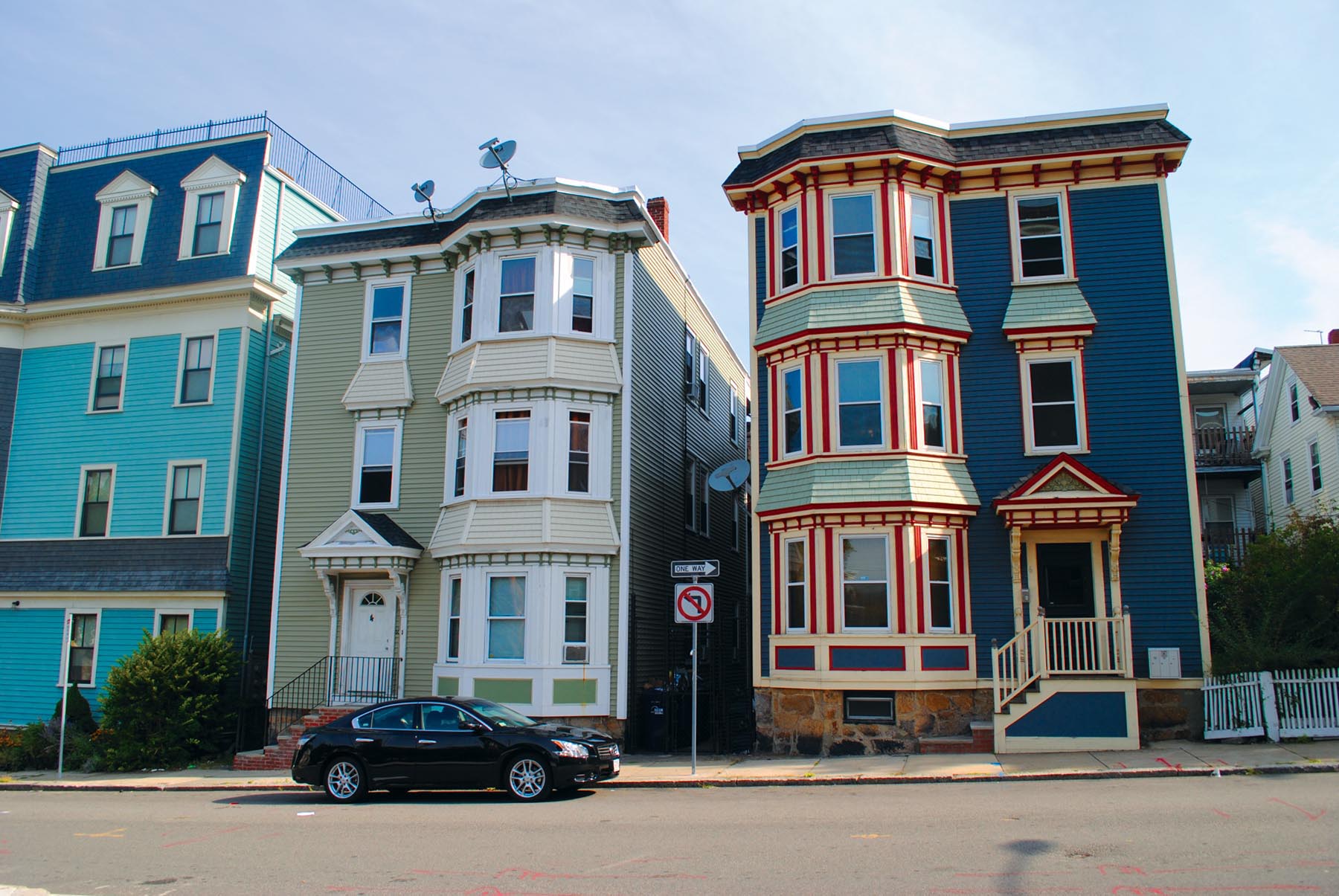 Photo is of two triple decker houses on a street. There is a car parked on the street in front of one of the houses.