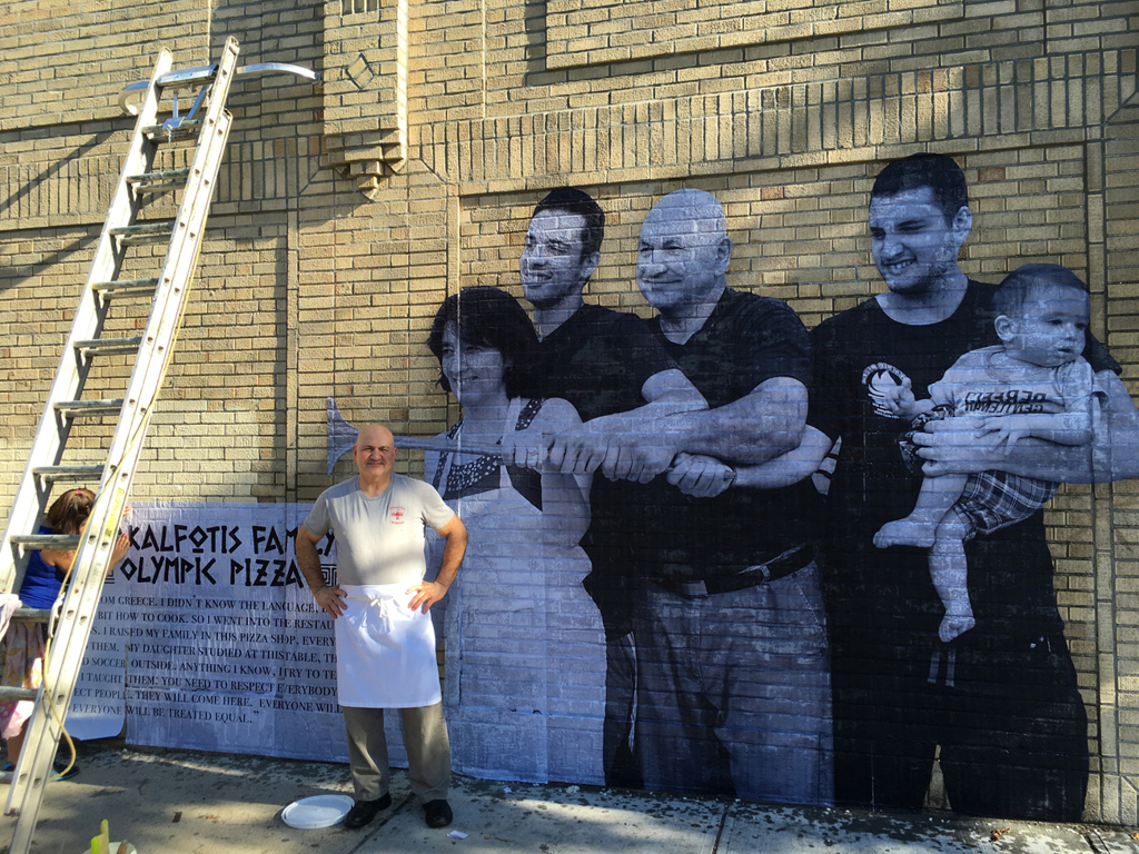 A man stands in front a mural of a family with "Kalfotis Family: Olympic Pizza" identified as the subjects