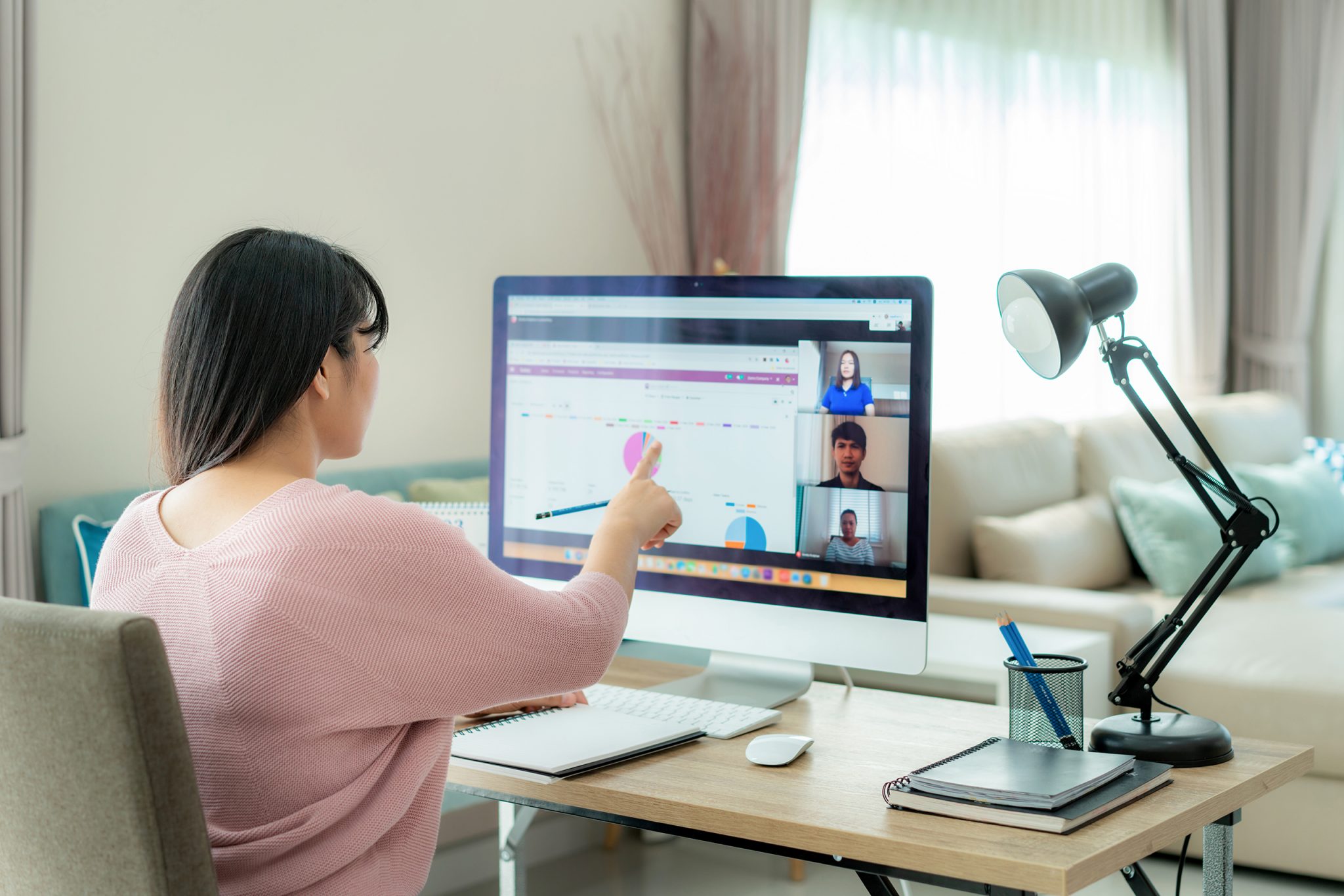 Asian business woman talking to her colleagues about plan in vid A woman with dark brown hair and who is wearing a pink sweater sits in front of a computer screen on a virtual call.