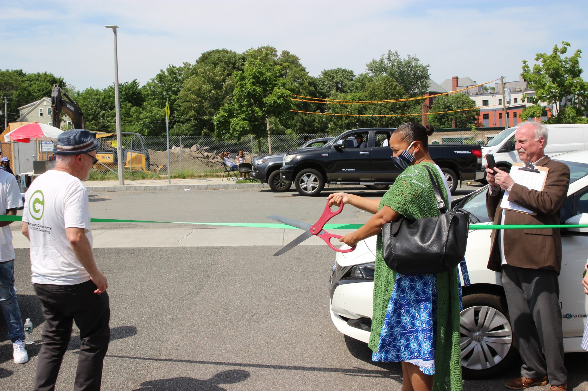 Reverend Mariama White-Hammond, Boston's Chief of Environment, uses a large pair of scissors to cut a green ribbon in front of a white vehicle.