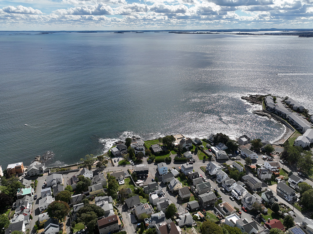 An aerial photo of the ocean in the top half of the photo and a blue sky and white clouds above the ocean. The bottom half of the photo is rows of homes and streets.
