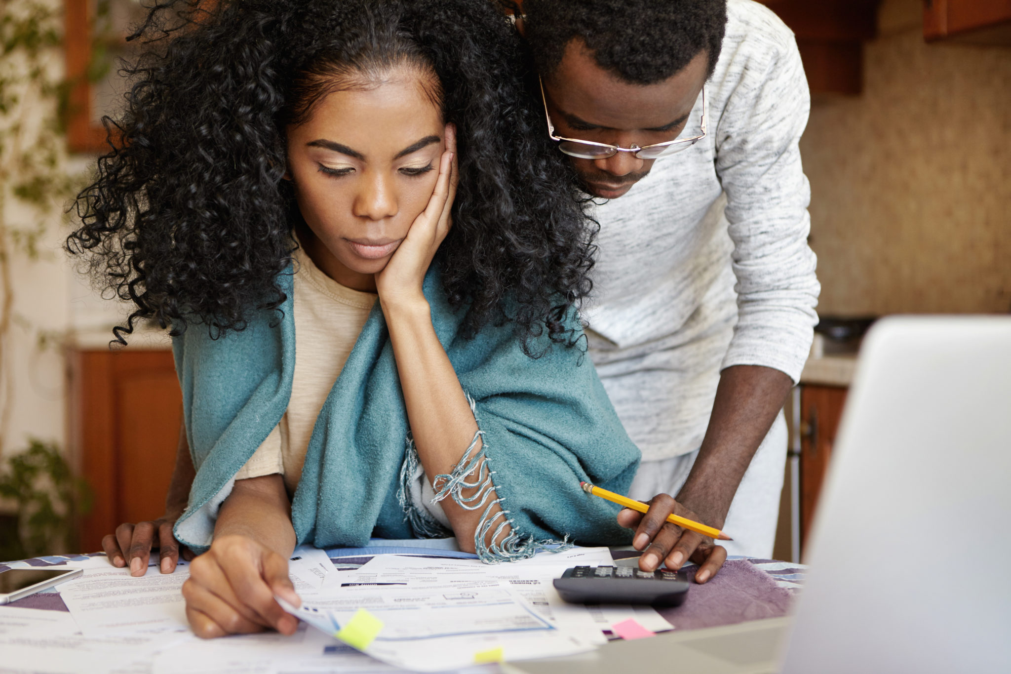 People, family budget, finances and payments concept. African-American man wearing glasses helping his frustrated wife doing paperwork, analyzing papers, calculating expenses, using calculator An African-American female sits at a table reviewing documents. She has an aqua colored blanket over her shoulders. An African-American male is leaning over her shoulder from behind reviewing the documents as well, and using a calculator.