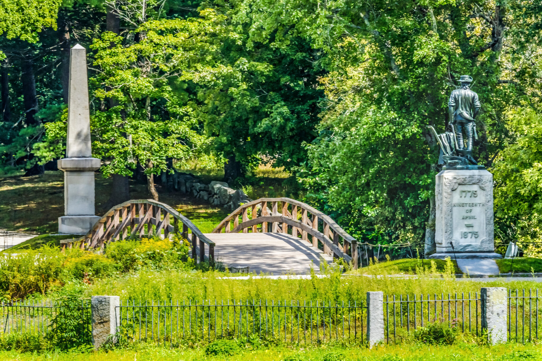 Photo is of the Minute Man Statue and the Old North Bridge in the Minute Man National Park. Both are surrounded by green grass and trees.