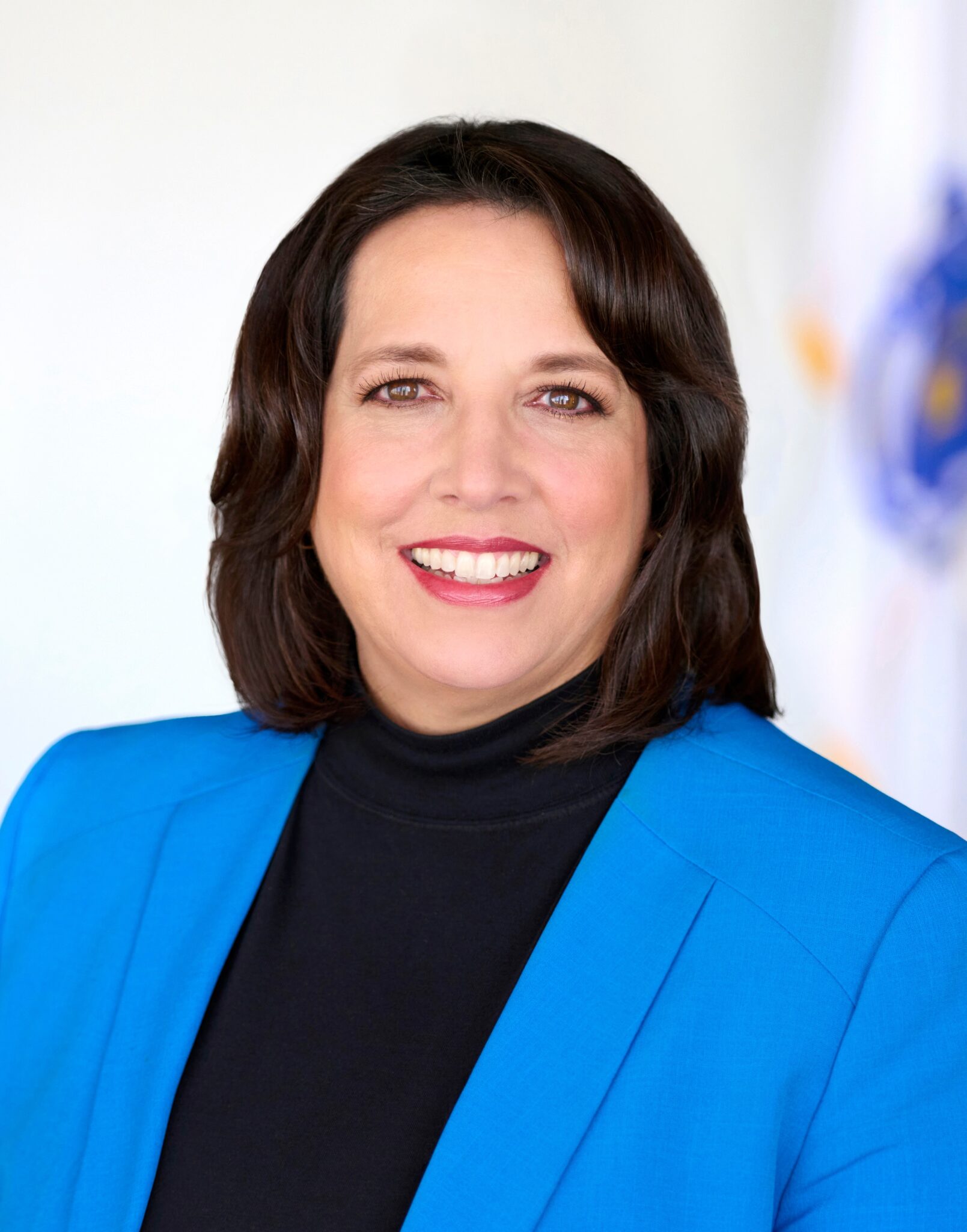 Headshot of Lt. Governor Kim Driscoll. She is wearing a black turtleneck with a blue blazer over it. She has medium-length brown hair, and she is smiling at the camera.