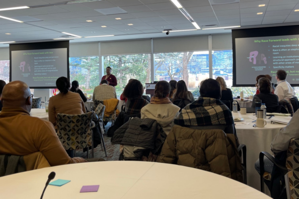 A room with people sitting in chairs at tables, facing the front of the room where someone is speaking behind a podium.