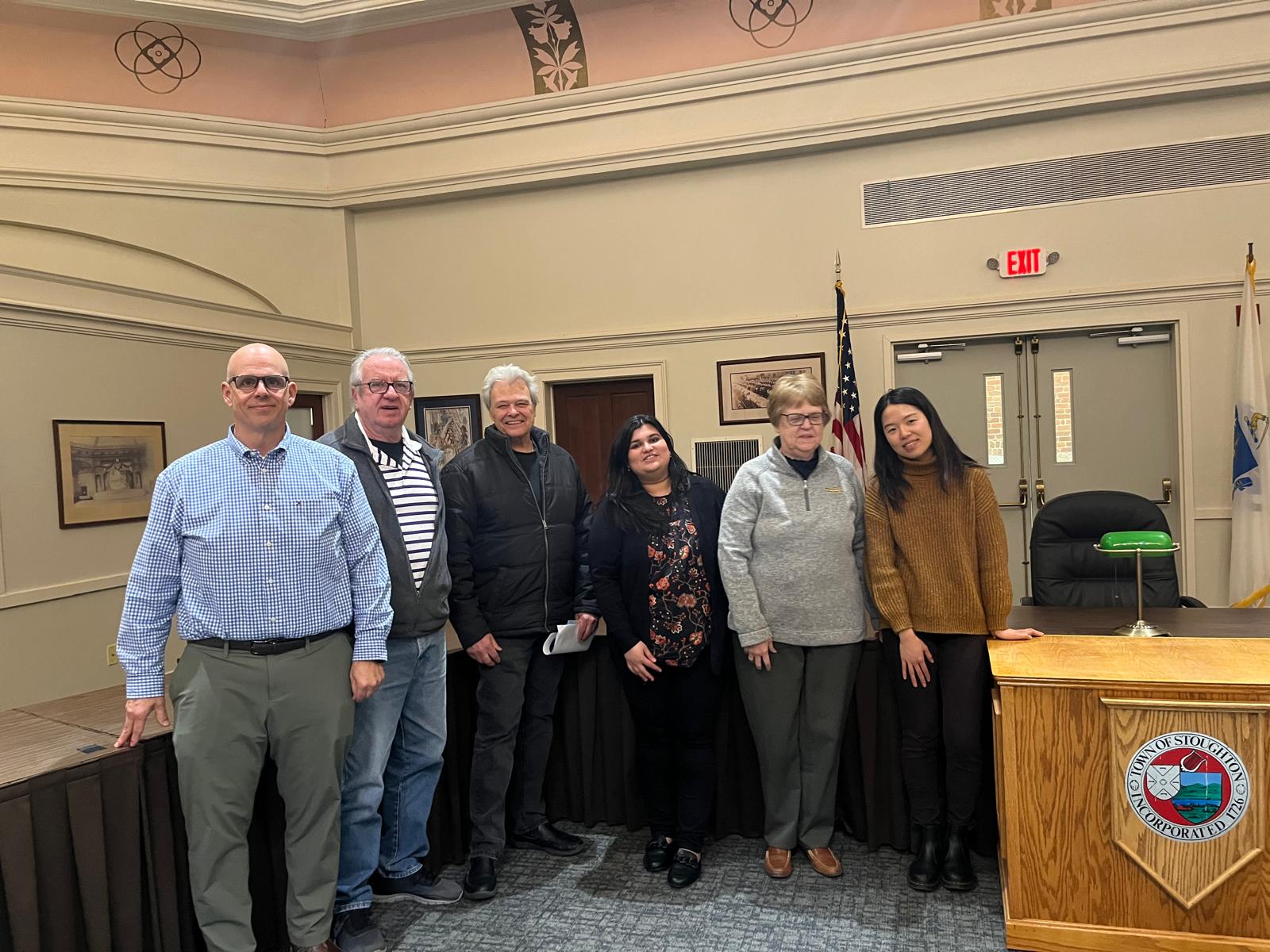 Route 27/Park Street Zoning Study Public Forum Event Six people stand together in a large room smiling at the camera. To the right of them is a desk with a black chair behind it.