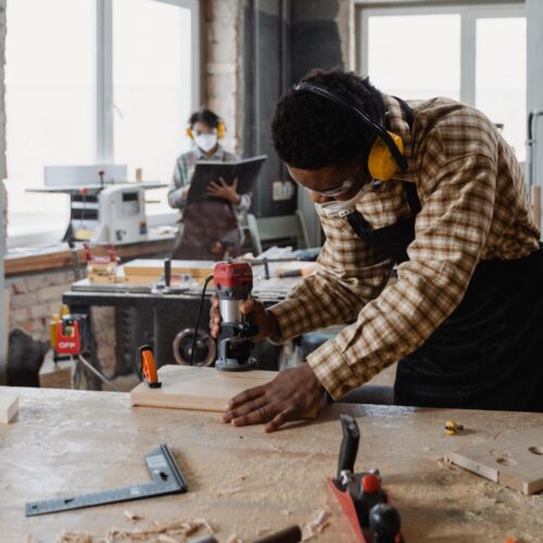 pexels-tima-miroshnichenko-6790977 An African-American man wearing a plaid shirt, apron, safety glasses, and safety ear muffs, stands over a work table using a tool on a piece of wood. Another person wearing a face mask stands in the background working at a work table.