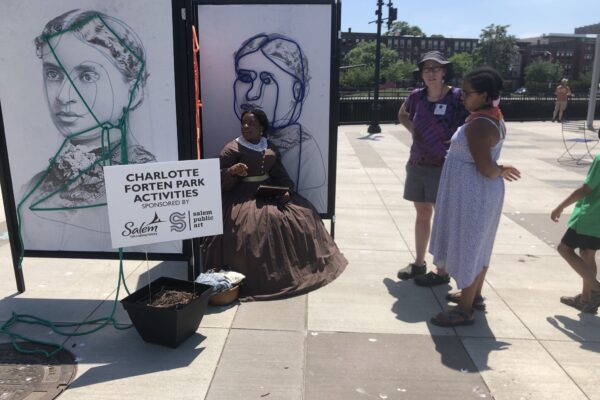 Two individuals are standing looking at artist renderings of two people on separate canvases with a woman sitting in front of one of them.