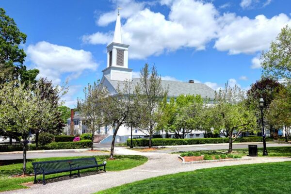 A park area with grass, trees, and walking paths. In the far background is a white church with a tall steeple.