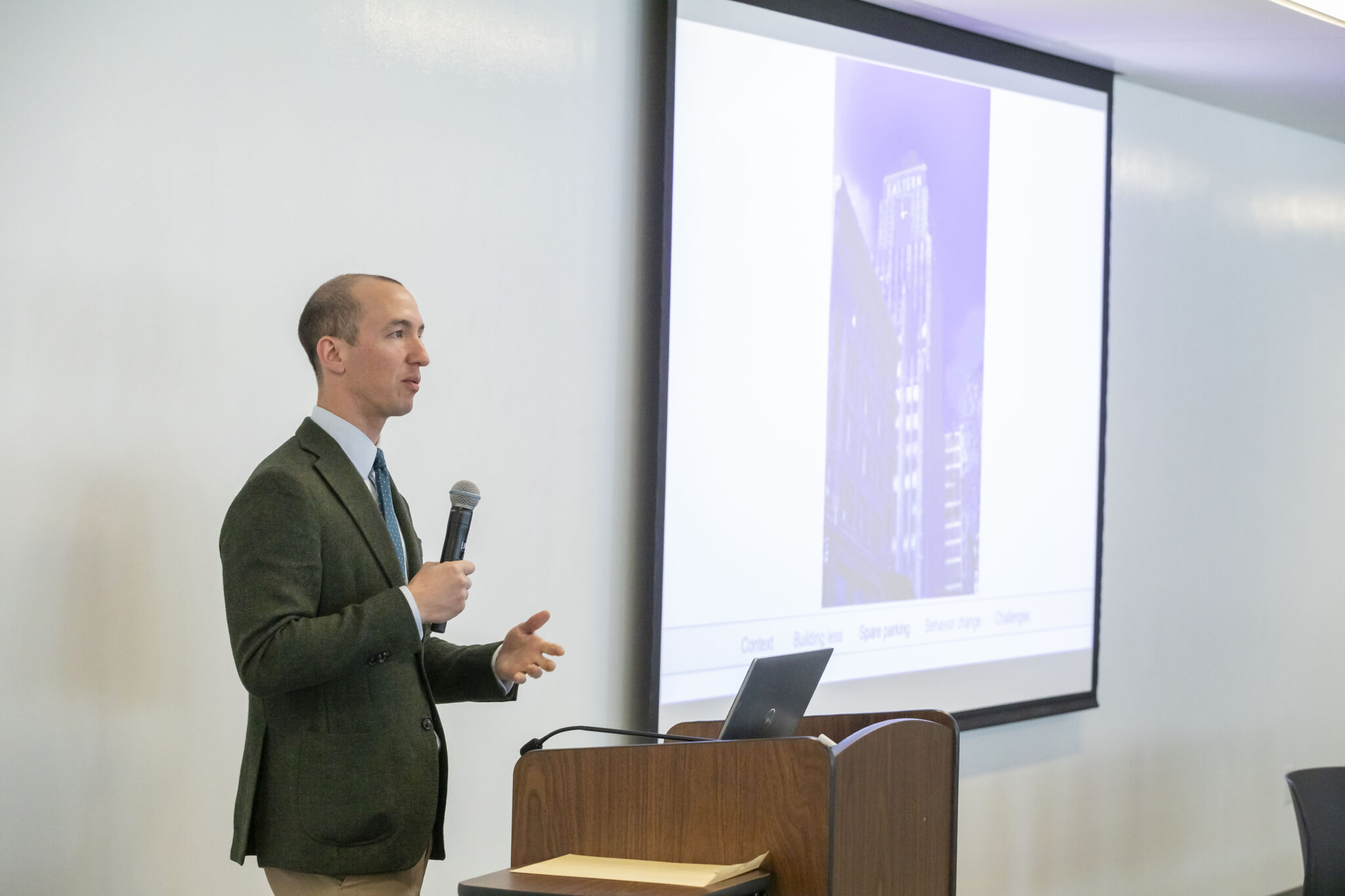 A close up of Henry Grabar speaking. He is standing behind a podium, speaking into a handheld microphone and to the right of him is a screen with a presentation on it.