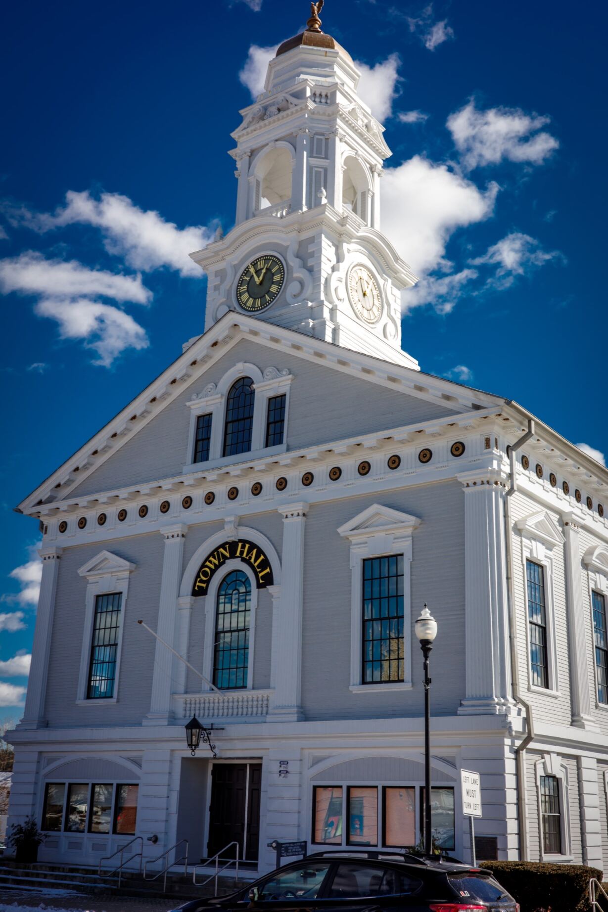 Photo is of the Milford Town Hall. A large building with a clock tower on the top. The sky is blue with a few white clouds above the building.