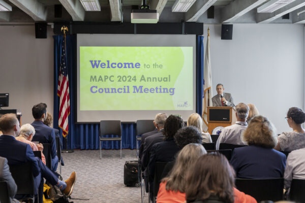 Photo shows people inside a conference room at the Commonwealth Museum - they are facing a screen and a speaker at a podium.