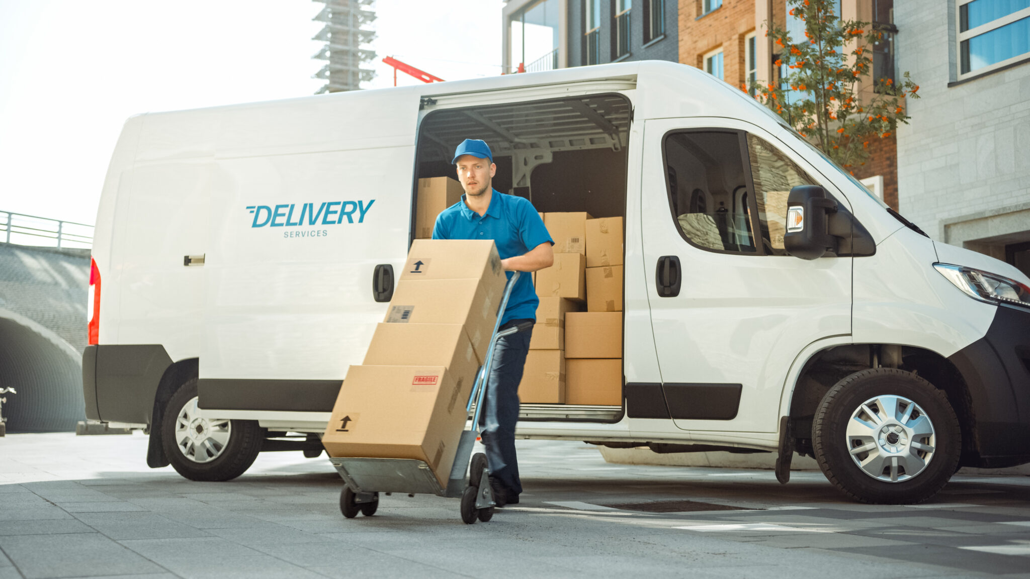 Delivery Man Pushes Hand Truck Trolley Full of Cardboard Boxes H A delivery person wearing a blue baseball cap, blue polo shirt, and black pants, is pushing a dolly with four stacked cardboard boxes on the dolly. Behind him is a white delivery van with the side door open showing stacks of cardboard boxes inside the van. blue writing on the door says, Delivery Services".