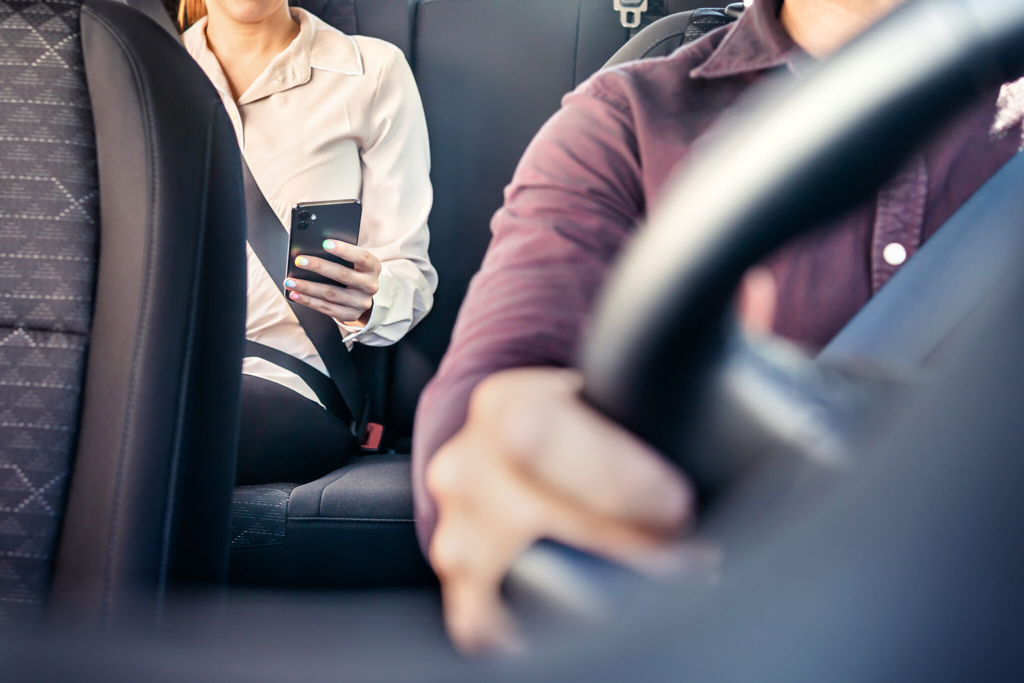 Phone in car or taxi. Passenger woman using cellphone in back seat of cab. Driver and customer. Rideshare mobile app. Professional business person travel to work, commute. Lady sitting in the backseat Female rider in a pink long sleeve shirt and black pants sits in the back of a ride share vehicle looking at her cell phone. The driver is wearing a dark purple top.