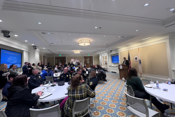 Photo of a conference room in the Massachusetts Statehouse. There are several tables with many law enforcement attendees, as well as a speaker standing at a podium.