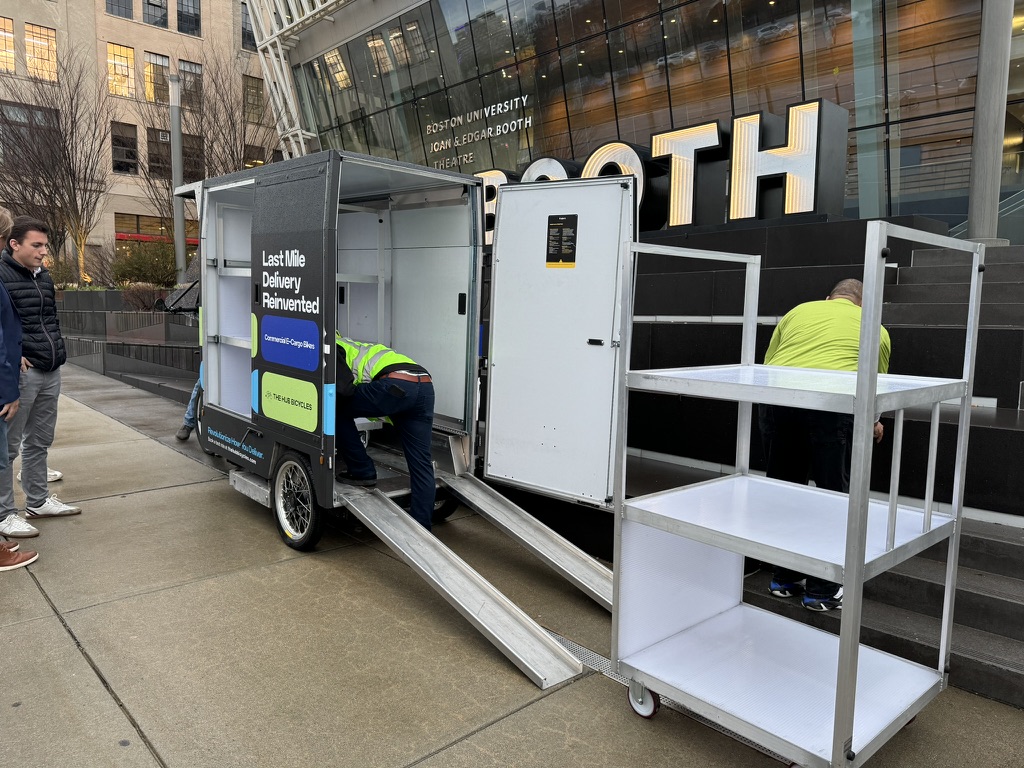 E-Cargo Bike in Boston2 Back of an E-Cargo bike, the cargo box is open and has two white ramps coming off of it. Behind the ramps is a three tiered white cart. There are two people with yellow vests on, one inside the cargo box, and one standing on the sidewalk next to it. There is a building behind the e-cargo bike, and two people standing to the left of it looking at it.