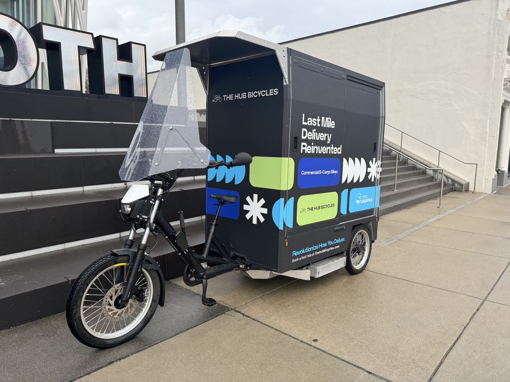Fulpra Cargo Trike - E-Cargo bikes webpage A black e-cargo bike with a black cargo box on the back of the bike. Numerous words and different colored text and design boxes are on the cargo box. It is parked on the sidewalk in front of stairs.