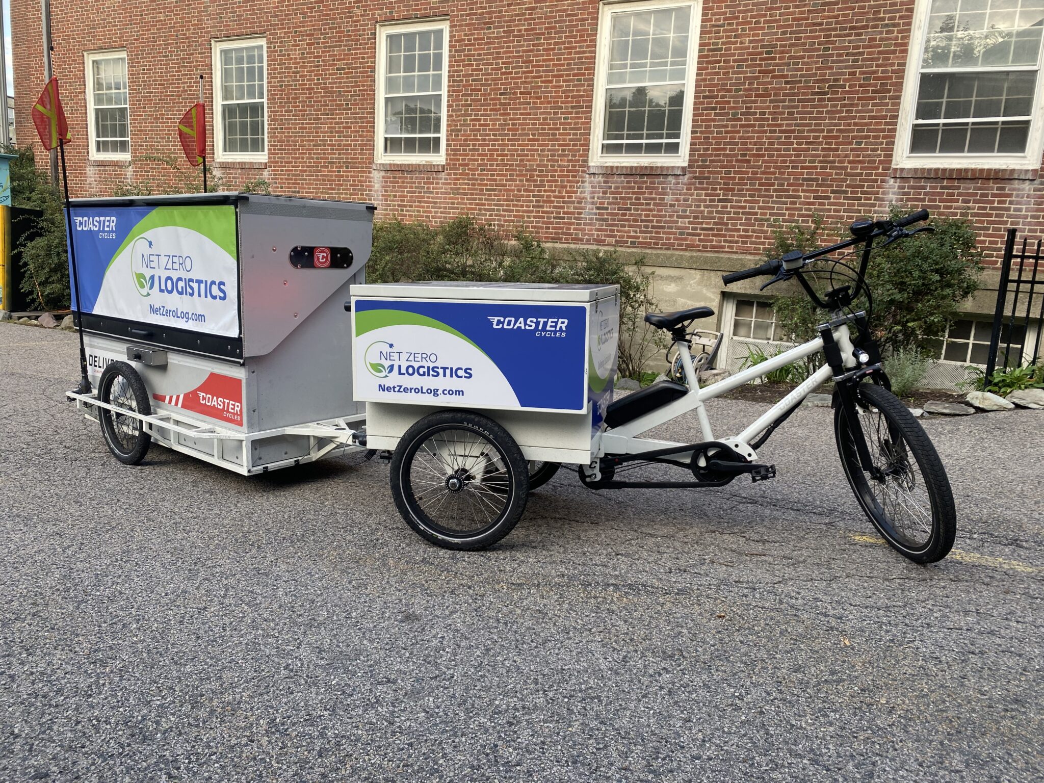 Coaster Cycle with Trailor - E-cargo bikes webpage A white e-cargo bike with a small cargo white and blue cargo box on the back with advertising text on it. On a trailor attached to the bike is another, larger multi colored cargo box with advertising box on it. The boke is parked next to a large brick building.