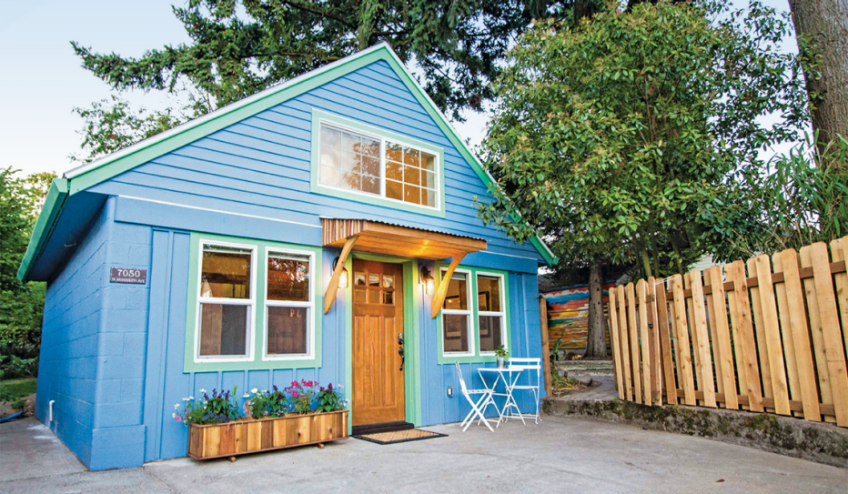 A small, blue, detached ADU. It has an orange front door, numerous windows, small planters in the front. It has trees and a wood fence in the background.
