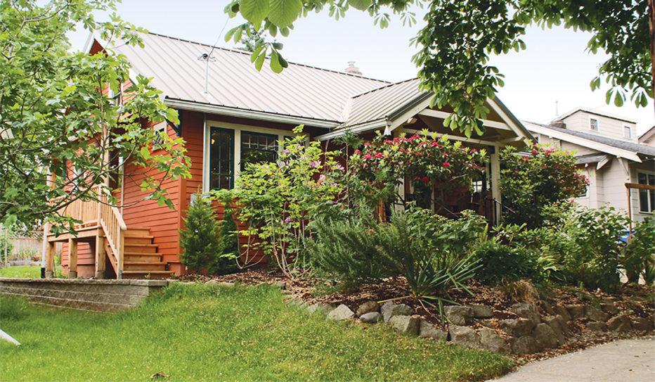 Internal ADU. An orange one level home. Stairs on the side of the home lead into the studio apartment. Stairs in the center of the home lead to the main entrance of the main home. There are rocks, plants, and grass surrounding the house.