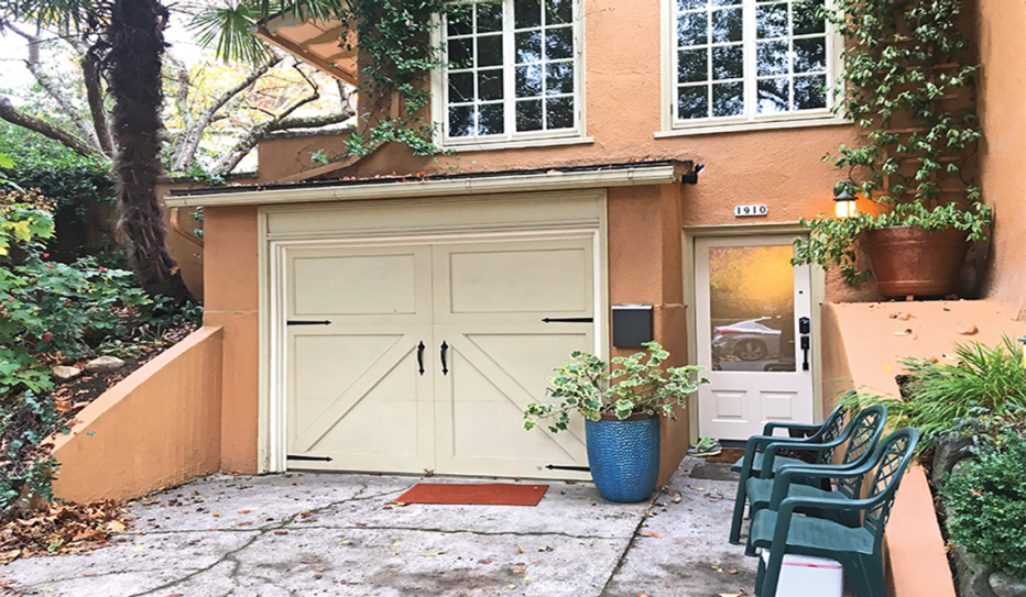 A lower level ADU. A peach stucco house with white window trim and doors. Bottom level of the home has vanilla colored garage doors that open from the front. There are three green patio chairs on the driveway, and plants and greenery surrounding the house.