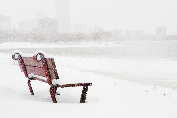 A snow covered red bench with snow all around it on the ground. The Charles River and Boston are barely visible in the background through the falling snow.