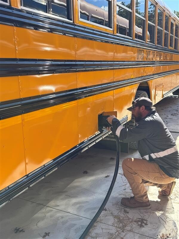 Image 1 - electric school bus webpage A person wearing construction type clothes kneels in front of the side of a school bus, holding and putting into the bus, an electric charging cable.