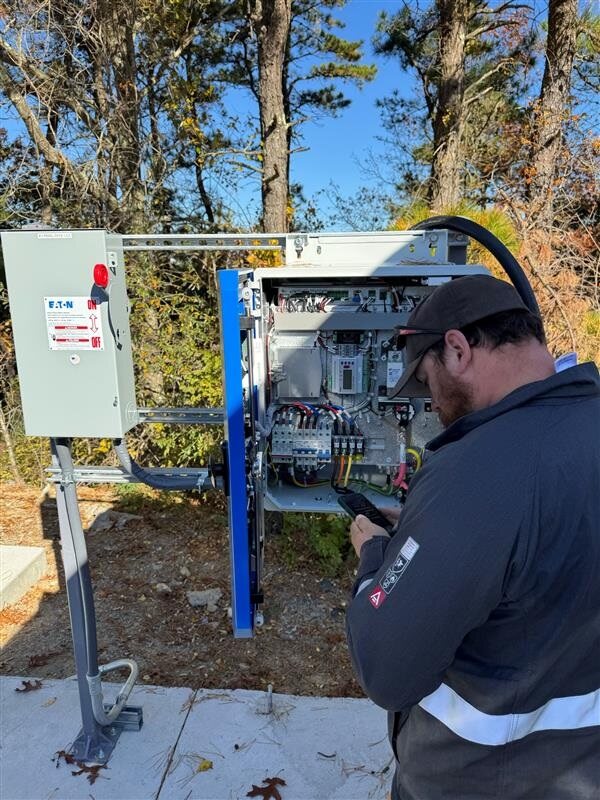 Image 4 - electric school bus webpage A person wearing green pants and a dark jacket with a beard and dark short hair, stands in front of an EV cable box, working on it.