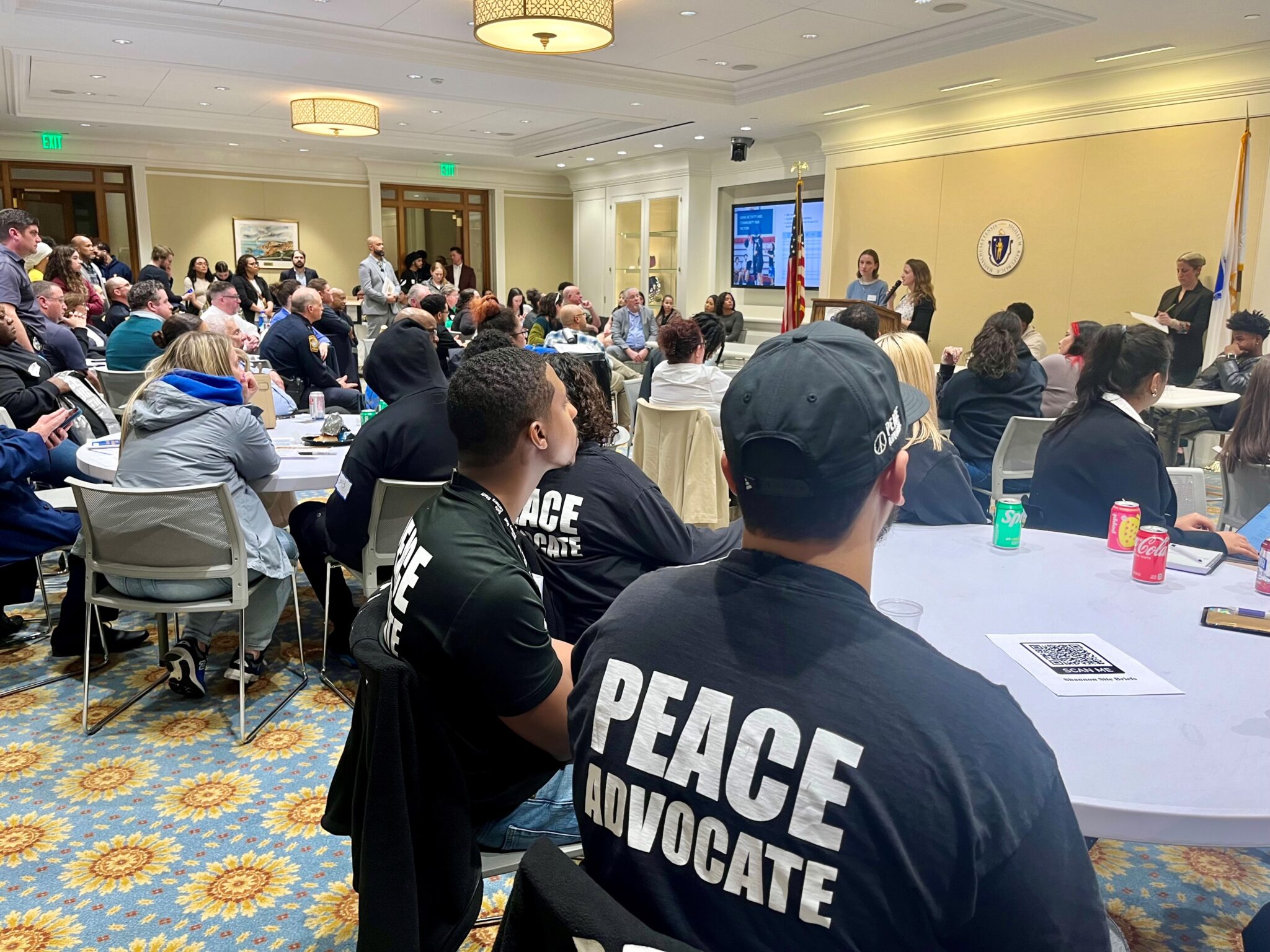 Two youth stand speaking into a microphone to people seated in chairs at white covered tables. In the forefront of the photo are two seated individuals wearing black tshirts with text in white letters on the back that says, "Peace Advocate".