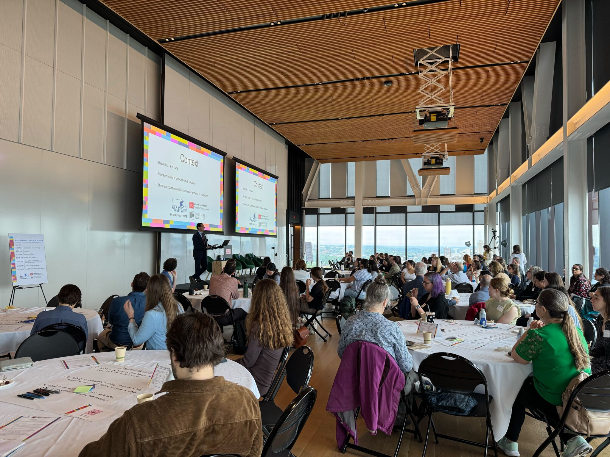A room full of people sitting at tables listening to someone speaking at the front of the room.