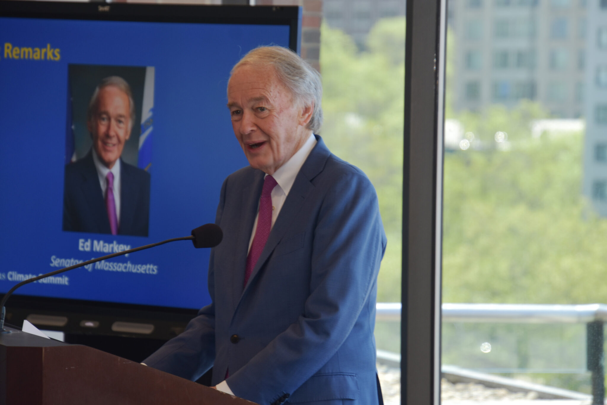 DSC_0795 U.S. Senator Edward Markey is standing behind a wooden podium speaking. To the right of him is a large projection screen with a photo of him, his name, and information on it.