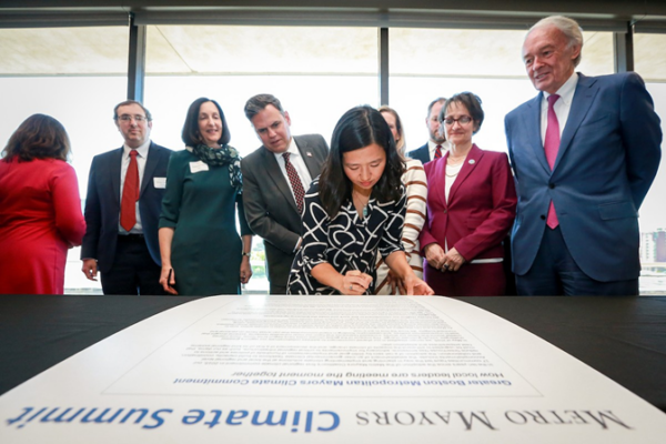 City of Boston Mayor Wu signs the 2025 Climate Commitment. Behind her, from left to right, are Melrose Mayor Jennifer Grigoraitis (facing away), Watertown Town Manager George Proakis, Newton Mayor Ruthann Fuller, Malden Mayor Gary Christenson, Medford Mayor Breanna Lungo-Kohen (partially hidden), Arlington Town Manager Jim Feeney (partially hidden), Mayor Katjana Ballantyne of Somerville, and Senator Ed Markey.