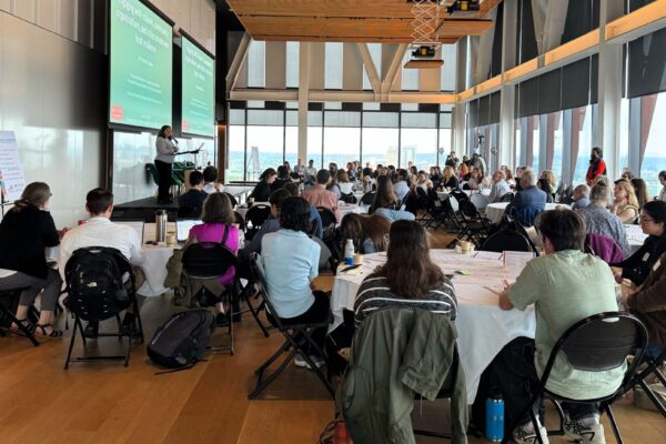 A view of the entire room at the Symposium to Advance Equitable Heat Health Actions. Attendees can be seen sitting at tables with a small stage and two projector screens on the left side of the room.