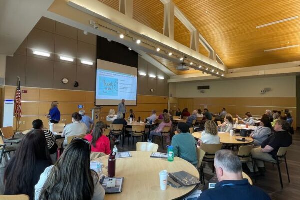A large room full of people sitting at round tables facing a speaker and a large projection screen with a presentation on it.