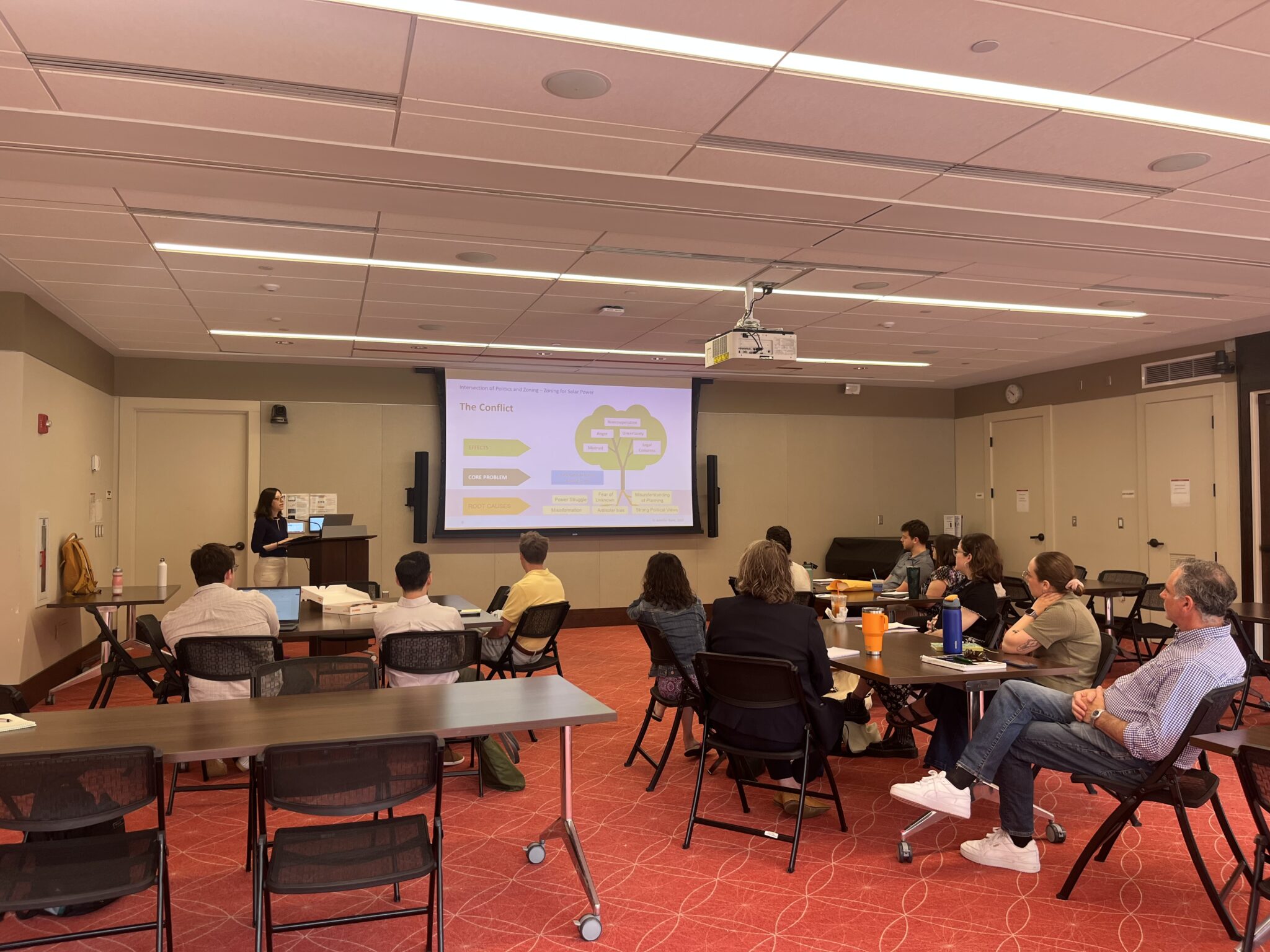NSPC June 2025 Meeting Volunteering Image A group of NSPC members sit in black chairs around several square tables, looking at the speaker in the foreground, standing behind a podium, a screen showcasing a slide from the presentation to their left.