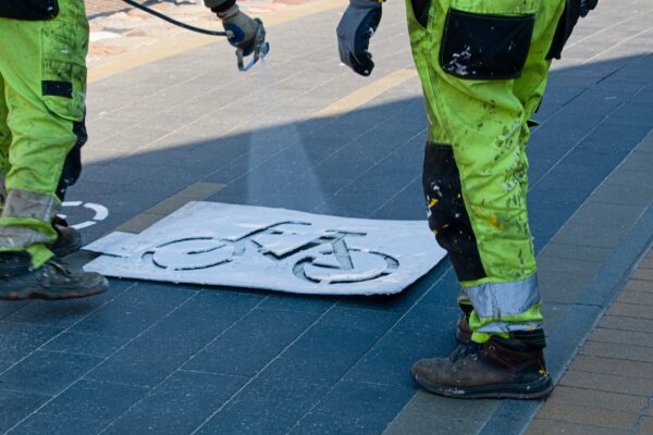 Bottom half (their pants and shoes) of workers painting a bike lane sign on a new bike lane.