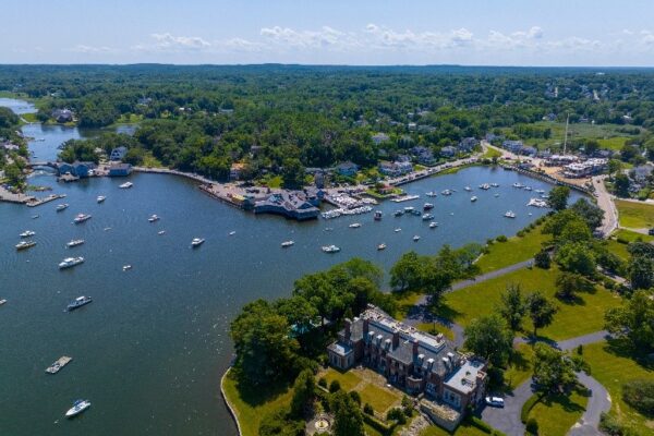 An aerial view of Cohasset Cove. An inlet can be seen with a dock and dozens of boats in the water. A large brick building is pictured in the foreground along the water.
