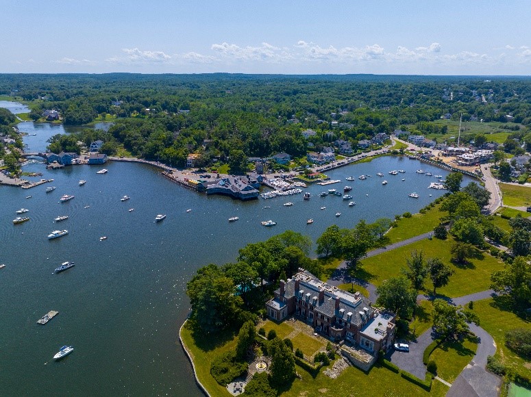 An aerial view of Cohasset Cove. An inlet can be seen with a dock and dozens of boats in the water. A large brick building is pictured in the foreground along the water.