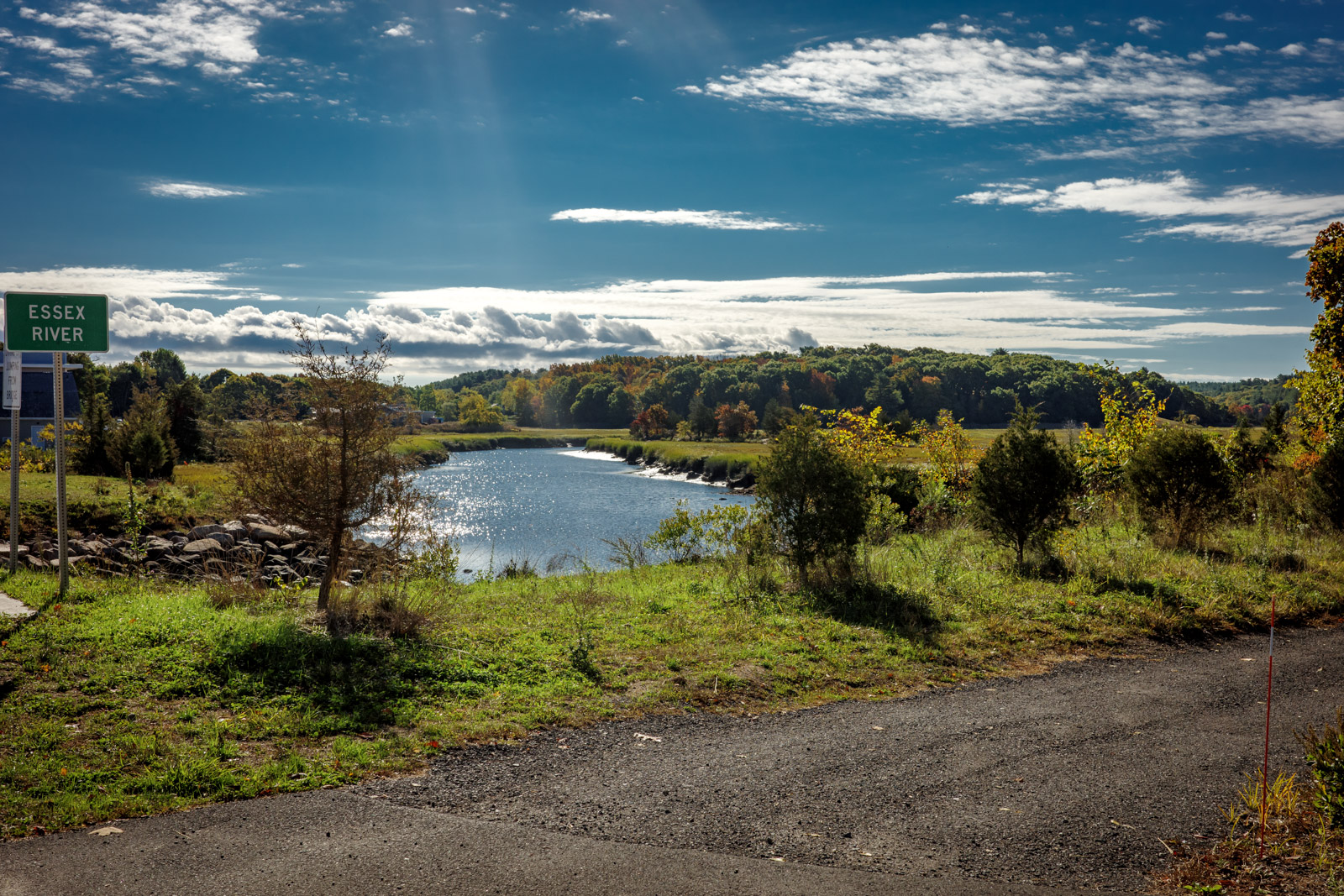 View of the Essex River from a walking path. A small brown sign says Essex River on it. A blue sky and slight clouds are over the river.