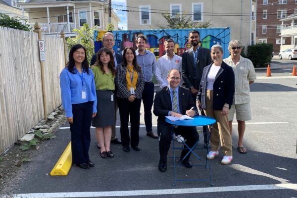 Mayor Dominick Pangallo is pictured at a small table with Lizzi Weyant to his left and a larger group of people standing behind. Photo was taken in a parking lot in Salem.