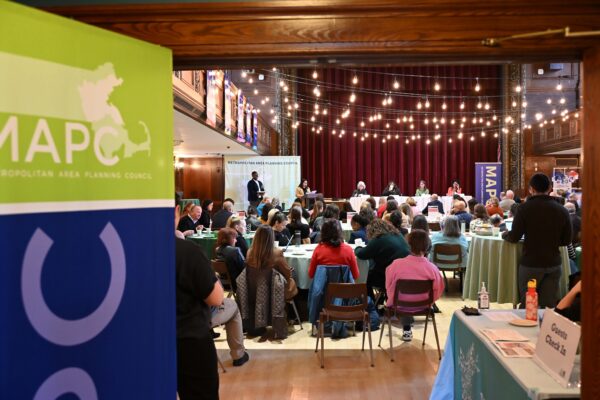A view looking into the Robbins Memorial Town Hall, with guests seated at tables and panelists seated towards a stage. A green and blue MAPC banner is pictured left close to the viewer.