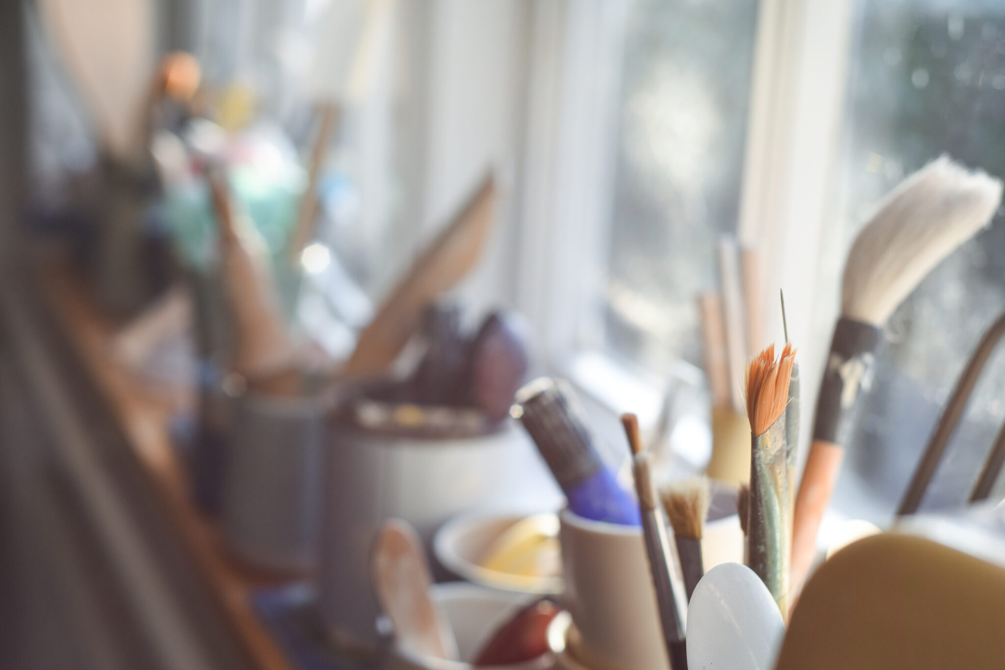 Cups of various arts supplies in them such as paintbrushes. The supplies are sitting on a window sill.