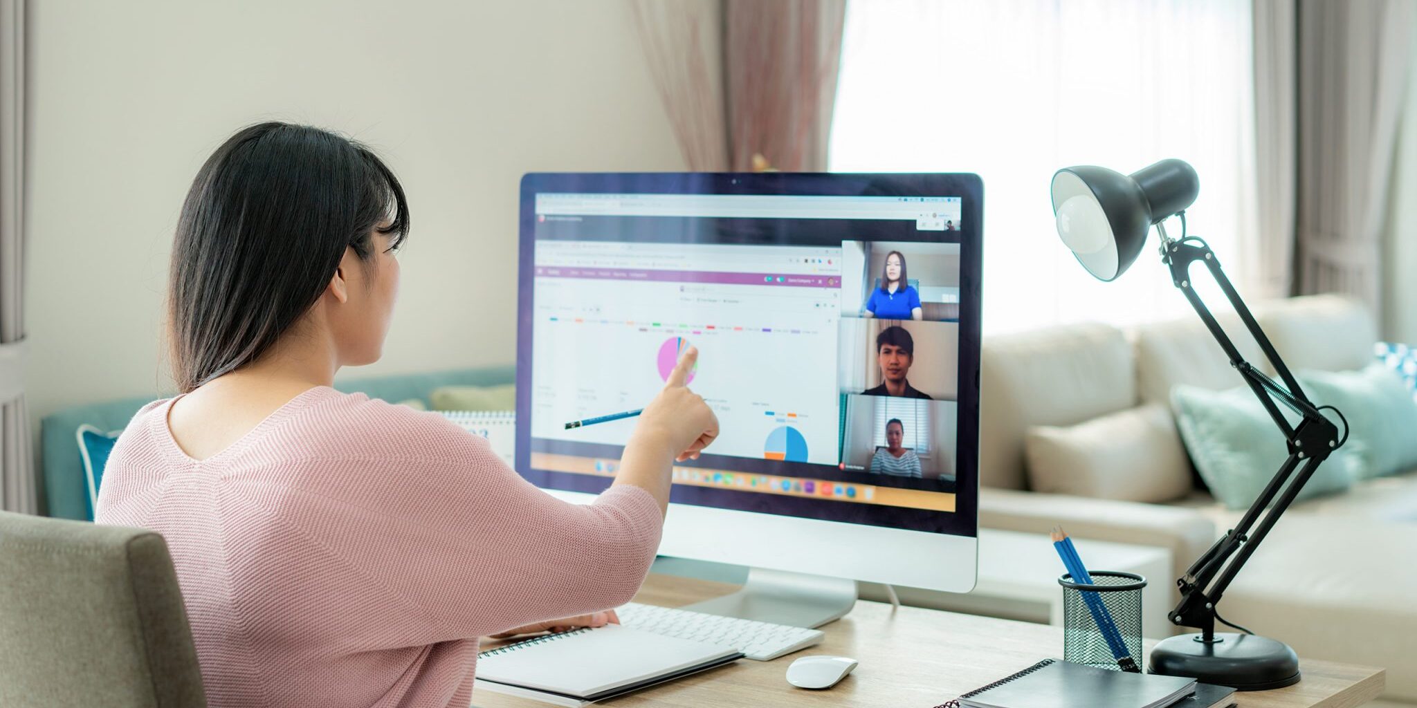 Asian business woman talking to her colleagues about plan in vid A woman with dark brown hair and who is wearing a pink sweater sits in front of a computer screen on a virtual call.
