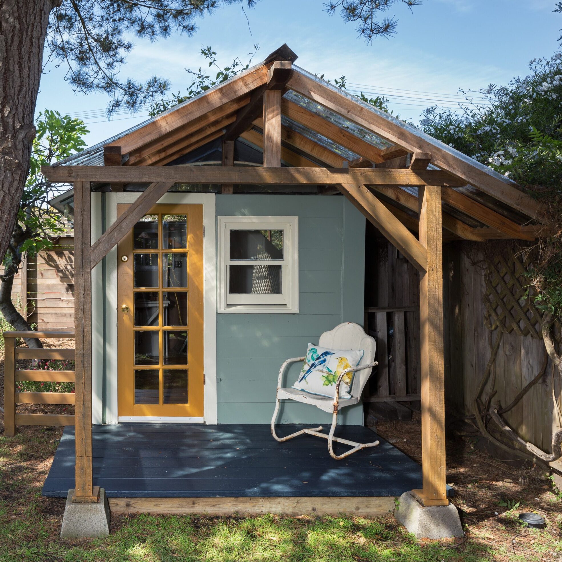 Cozy country style guest house Small guest house in light blue with a white trimmed window, a wooden front door with mini windows, a wood awning, and a blue floored front porch with a white chair with a flowered pillow siting on the front porch.