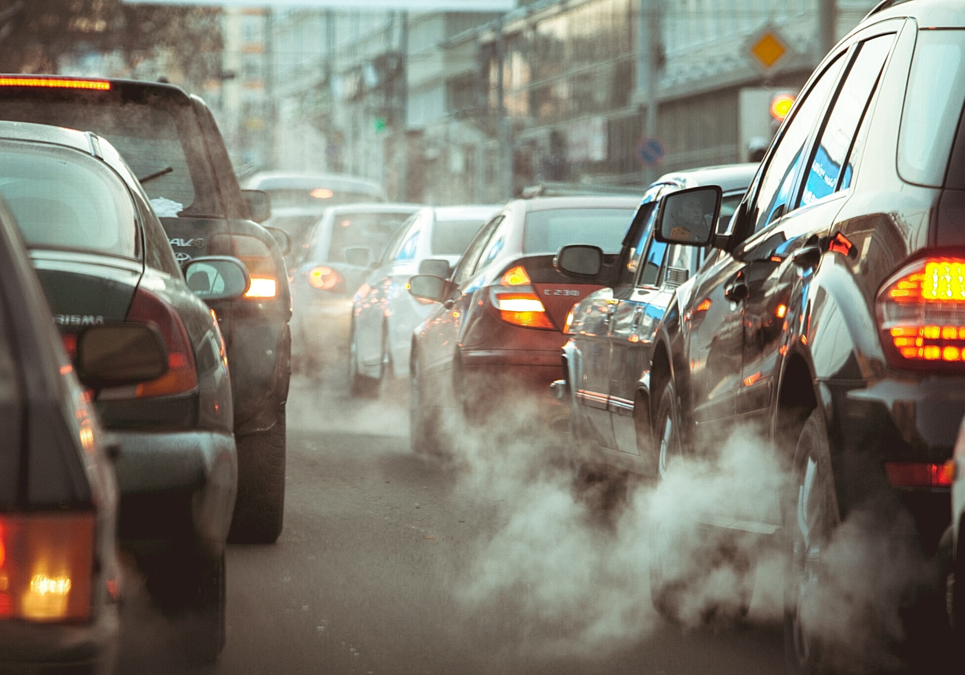 View from behind of many cars driving down the road while their exhaust pipes are emitting smoke or pollutants.
