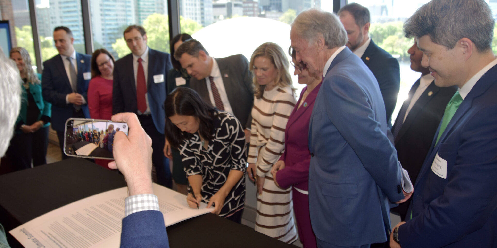 Boston Mayor Wu leans over the 2025 Climate Commitment signing it. MMC members, MAPC staff, Senator Markey, and others stand around her.