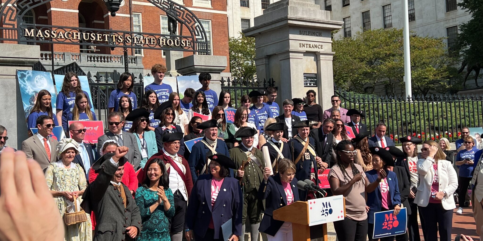 IMG_5954 Boston Mayor Wu, LG Driscoll, and Governor Healey stand with numerous other people, some dressed in revolutionary attire in front of the State House. Governor Healey is speaking at a podium.